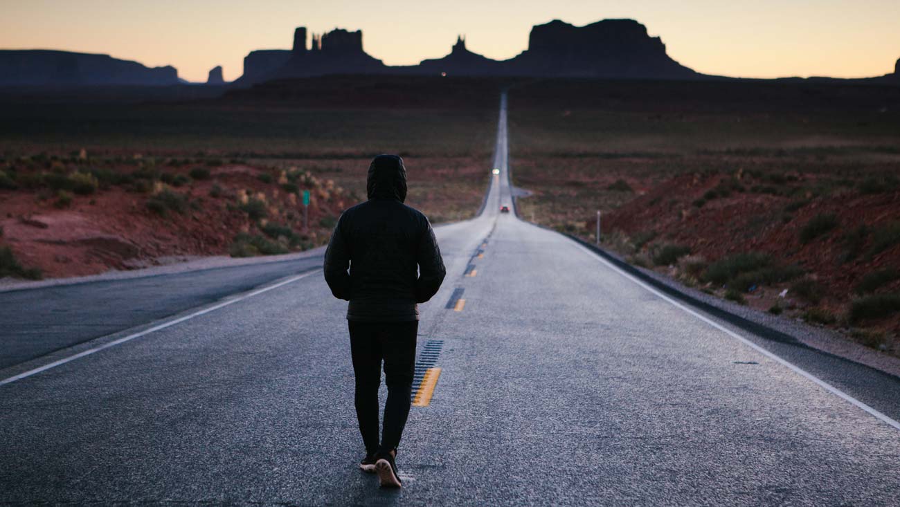 ragazzo visto da dietro in tenuta sportiva con felpa e cappuccio cammina lungo una strada asfaltata vuota