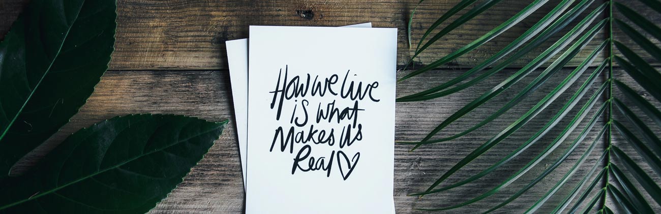 Biglietto bianco con scritto "How we live is what makes us real" su un tavolo di legno con foglie verdi di una palma accanto.